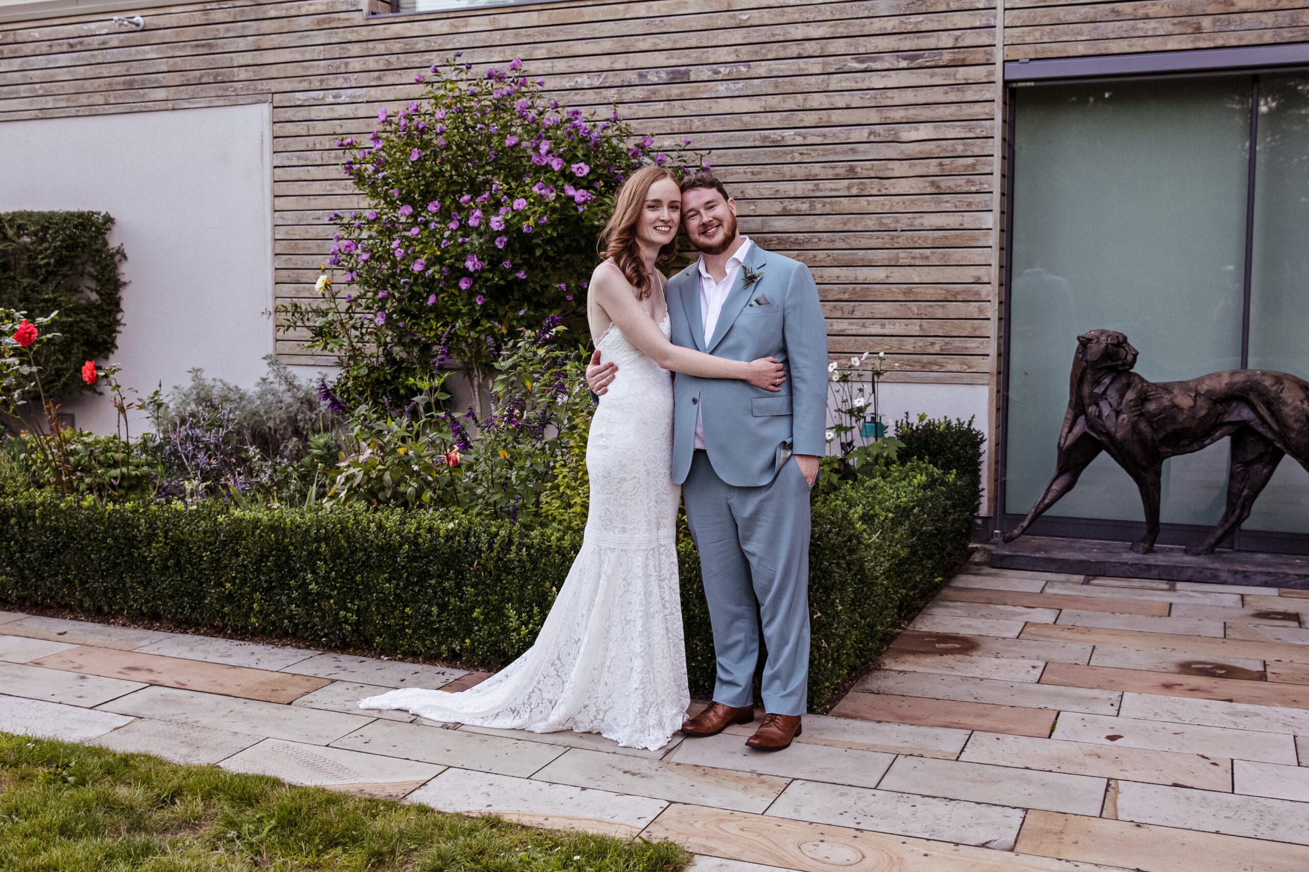 the bride and groom pose for picture in their garden with sculpture in background