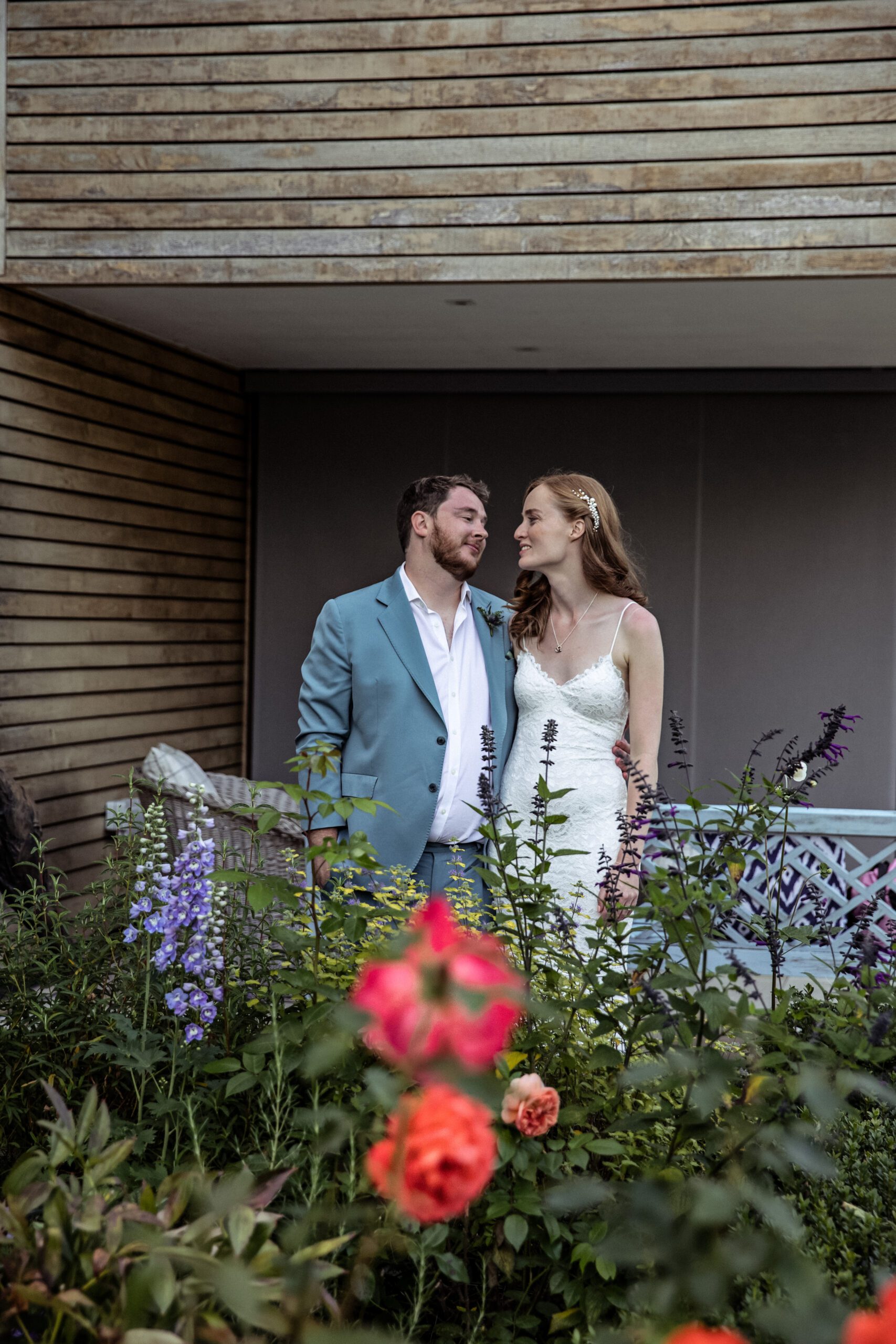 the bride and groom pose for picture in their garden with flowers in foreground