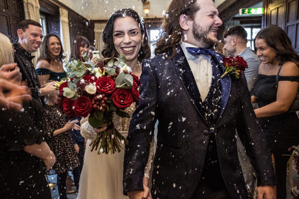 A bride and groom laugh as confetti is thrown in their direction by guests