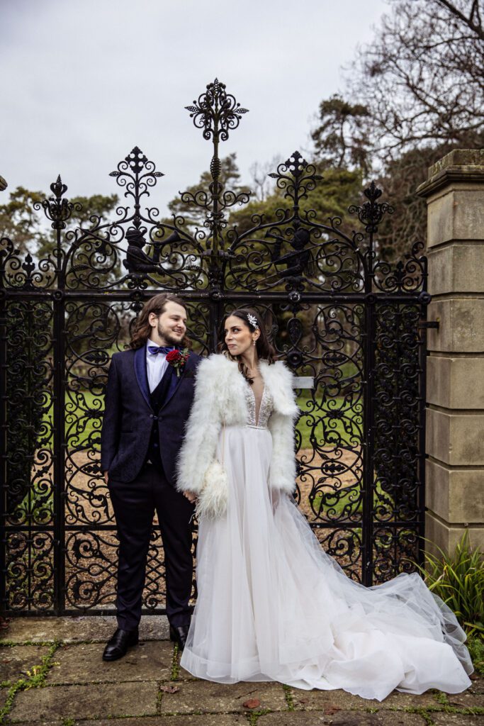 A bride and groom poses at the iron gates of Fanhams Hall gardens. They both hold hands and look at each other.
