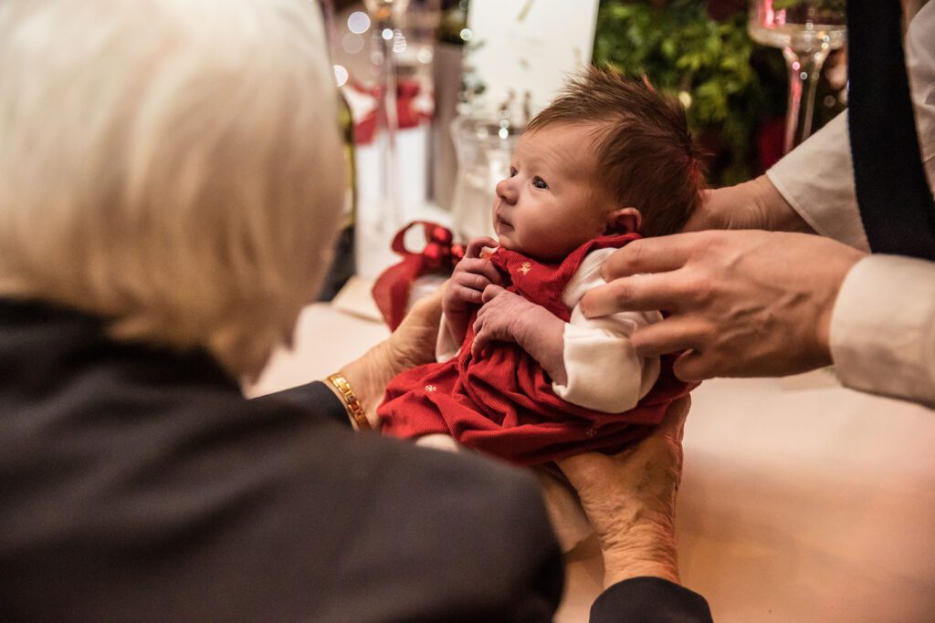 A newborn baby attends a wedding at Fanhams Hall. Her great Grandma is holding her. She is wearing a red dress